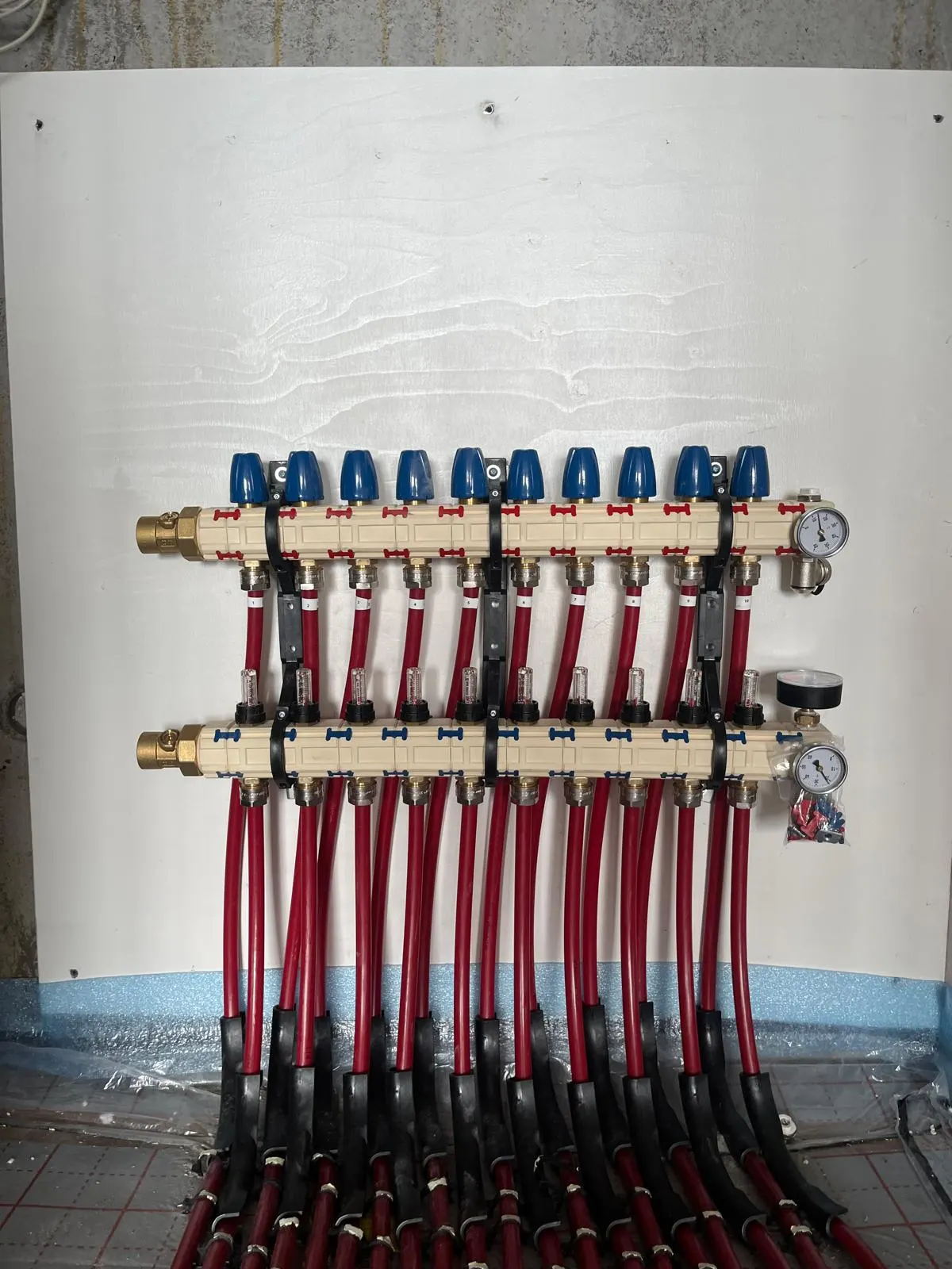 Een verdeler met tien groepen op een witte montageplaat tegen een betonwand, met blauwe afsluiters bovenop, doorstroommeters in het midden en rode leidingen die in zwarte bochtbeschermers naar de vloer met tackerfolie lopen