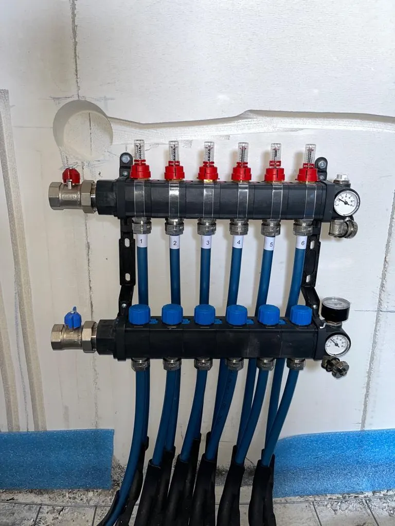 Een verdeler met zes groepen op een witte isolatieplaat, met bovenin de doorstroommeters met rode kappen en onderin de blauwe afsluiters, de aanvoerleidingen genummerd van 1 tot en met 6 en twee manometers ernaast, met blauwe randisolatie langs de vloer