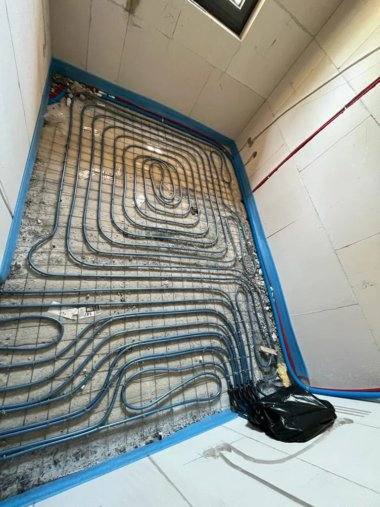 Een kleine ruimte helemaal omkaderd door blauwe randisolatie, met in het midden een slakkenhuis en daaromheen een serpentine van blauwe vloerverwarmingsbuis op een wapeningsnet, en rechts een wand van kalkzandsteen met een rode en een blauwe leiding erlangs