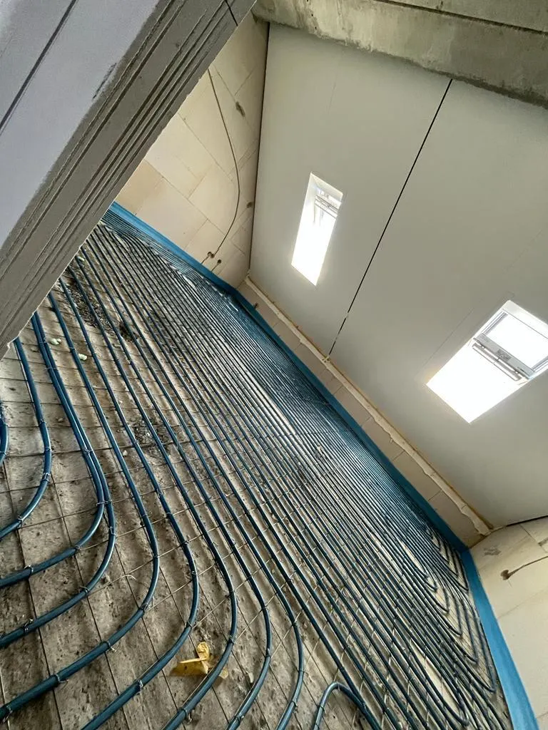 Een vertrek onder een schuine kap met twee dakramen erin, met blauwe vloerverwarmingsbuis in dichte rijen over de vloer op een wapeningsnet en een betonnen rand op de voorgrond