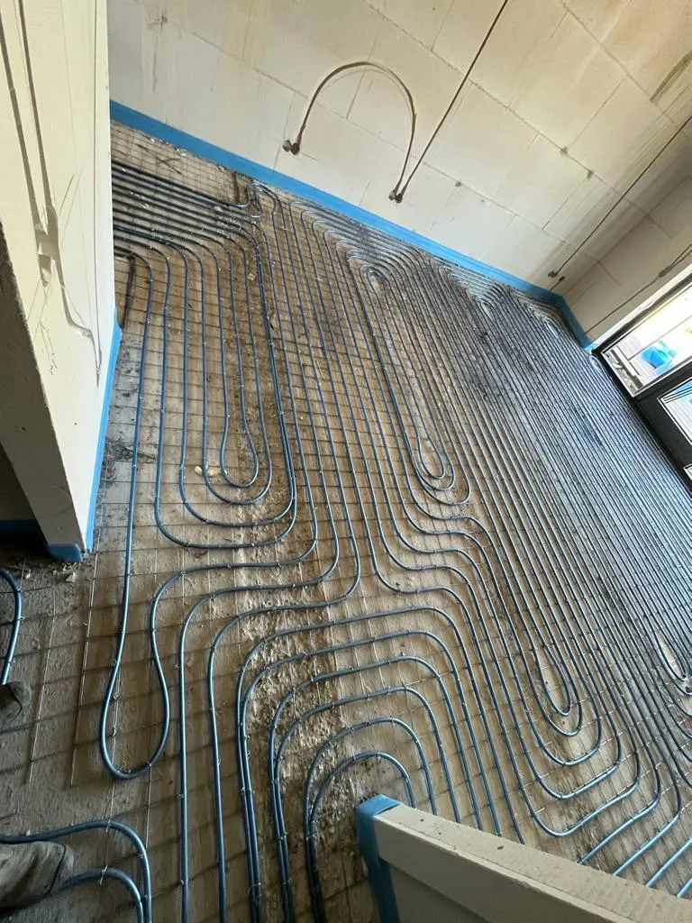 Een ruimte met blauwe vloerverwarmingsbuis in dichte lussen en ruime bochten op een wapeningsnet over een ruwe ondergrond, met blauwe randisolatie langs de wand, een raam rechts, een doorgang links en een losse elektraleiding die in een lus aan de wand hangt