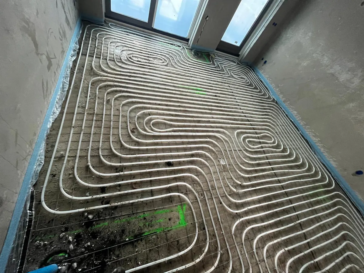 Een hele kamervloer vol vloerverwarmingsbuis in strakke lussen op een wapeningsnet, met blauwe randisolatie langs de wanden en een glazen pui met daglicht aan de kopse kant