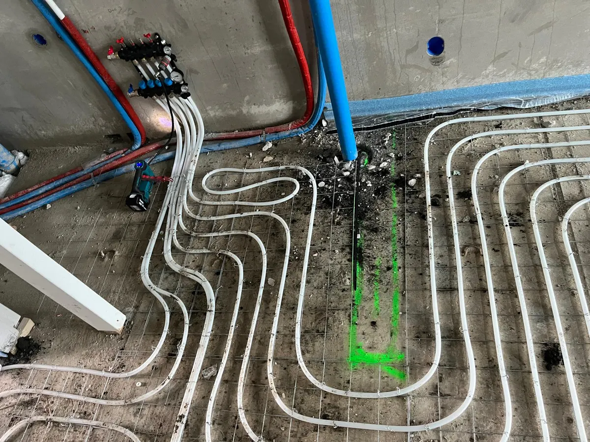 Witte vloerverwarmingsbuis die vanaf een verdeler aan de wand uitwaaiert over een wapeningsnet, met rode en blauwe geïsoleerde aanvoerleidingen langs de wand en een groene markering op de betonvloer