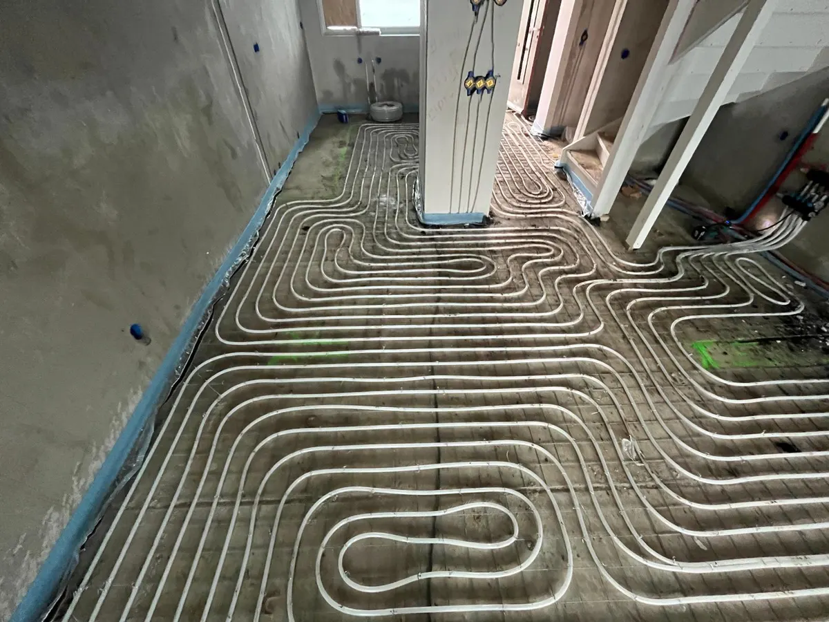 Een woonkamervloer vol vloerverwarmingsbuis op een wapeningsnet die om een leidingschacht met elektrabuizen heen loopt, met een houten trapopgang en een verdeler tegen de wand rechts