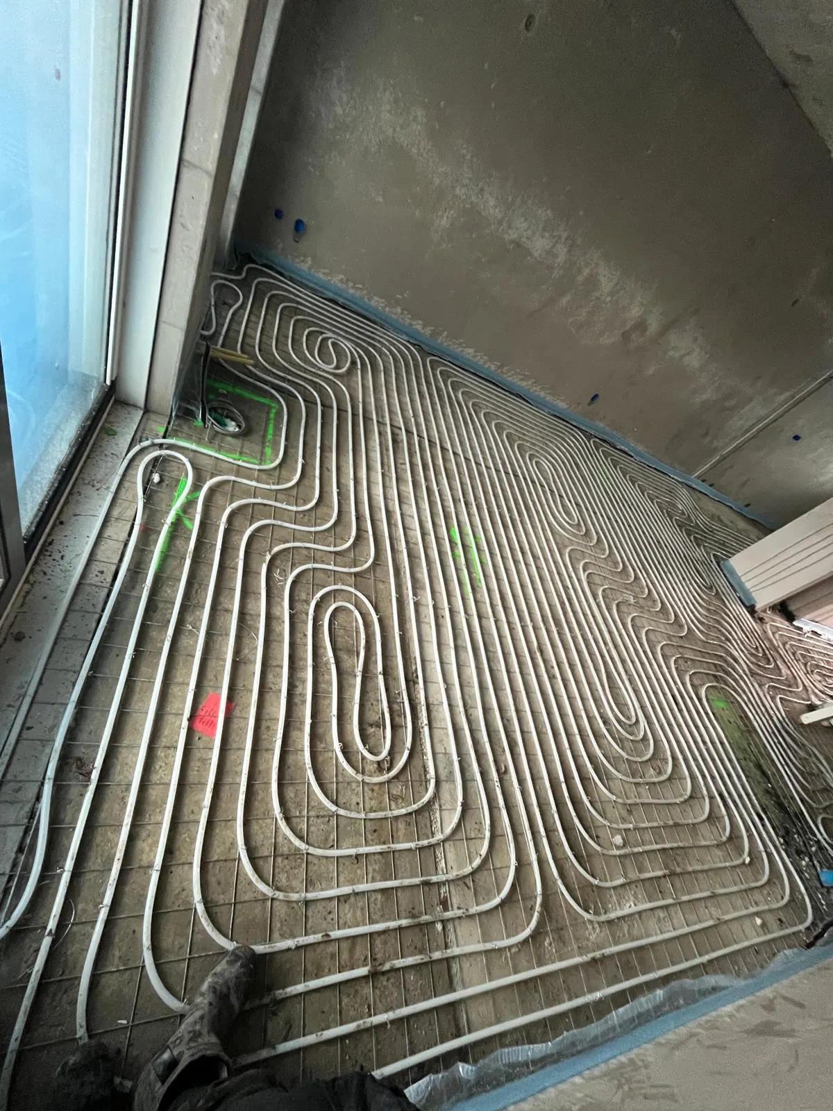 Vloerverwarmingsbuis in ineengeschoven lussen op een wapeningsnet langs een raampui, met groene en rode markeringen op de betonvloer en bouwmateriaal langs de wand