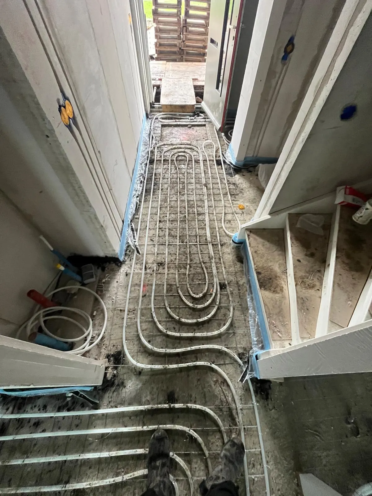 Vloerverwarmingsbuis in een lange lus op een wapeningsnet door een gang naar een open voordeur, met een rol buis en gereedschap langs de wand en houten stijlen aan de andere kant