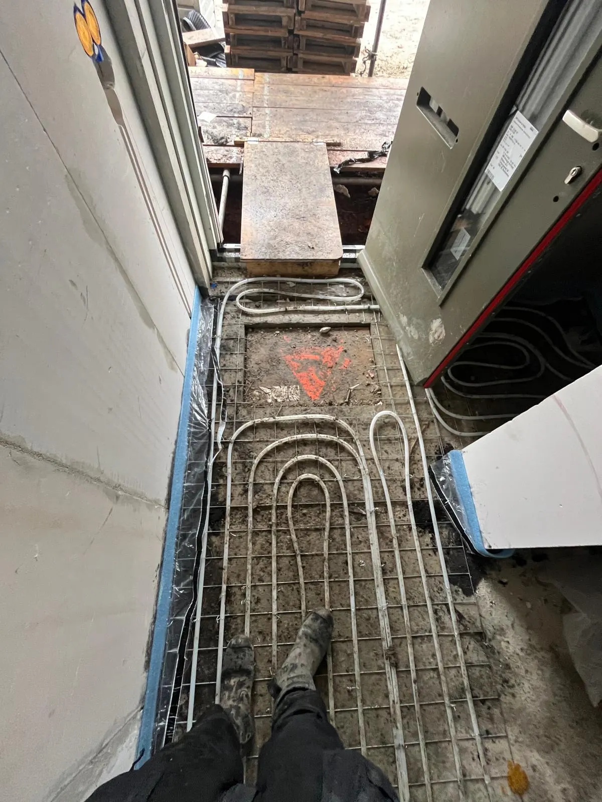 Vloerverwarmingsbuis in strakke bochten op een wapeningsnet bij een voordeur, langs een grijze meterkast, met een oranje markering op de betonvloer