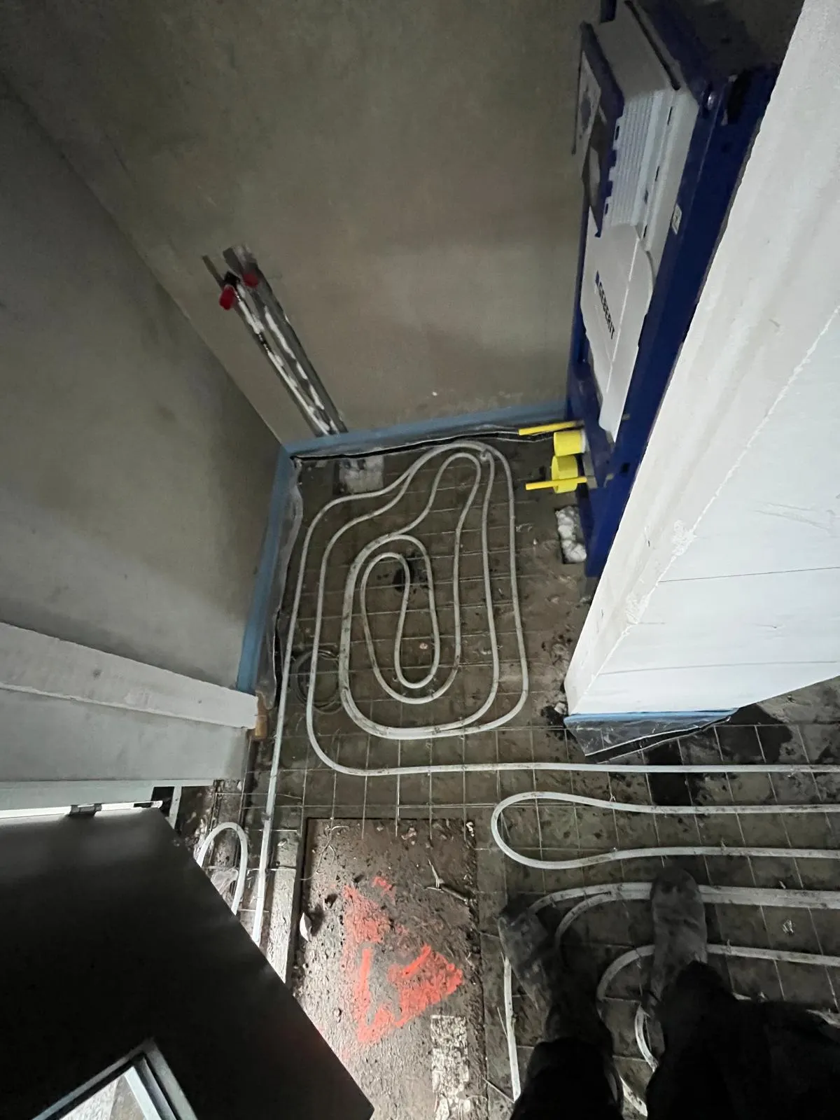 Vloerverwarmingsbuis in een enkele lus op een wapeningsnet in een kleine ruimte, met een blauw inbouwreservoir voor een hangend toilet tegen de wand en een aluminium ladder erachter