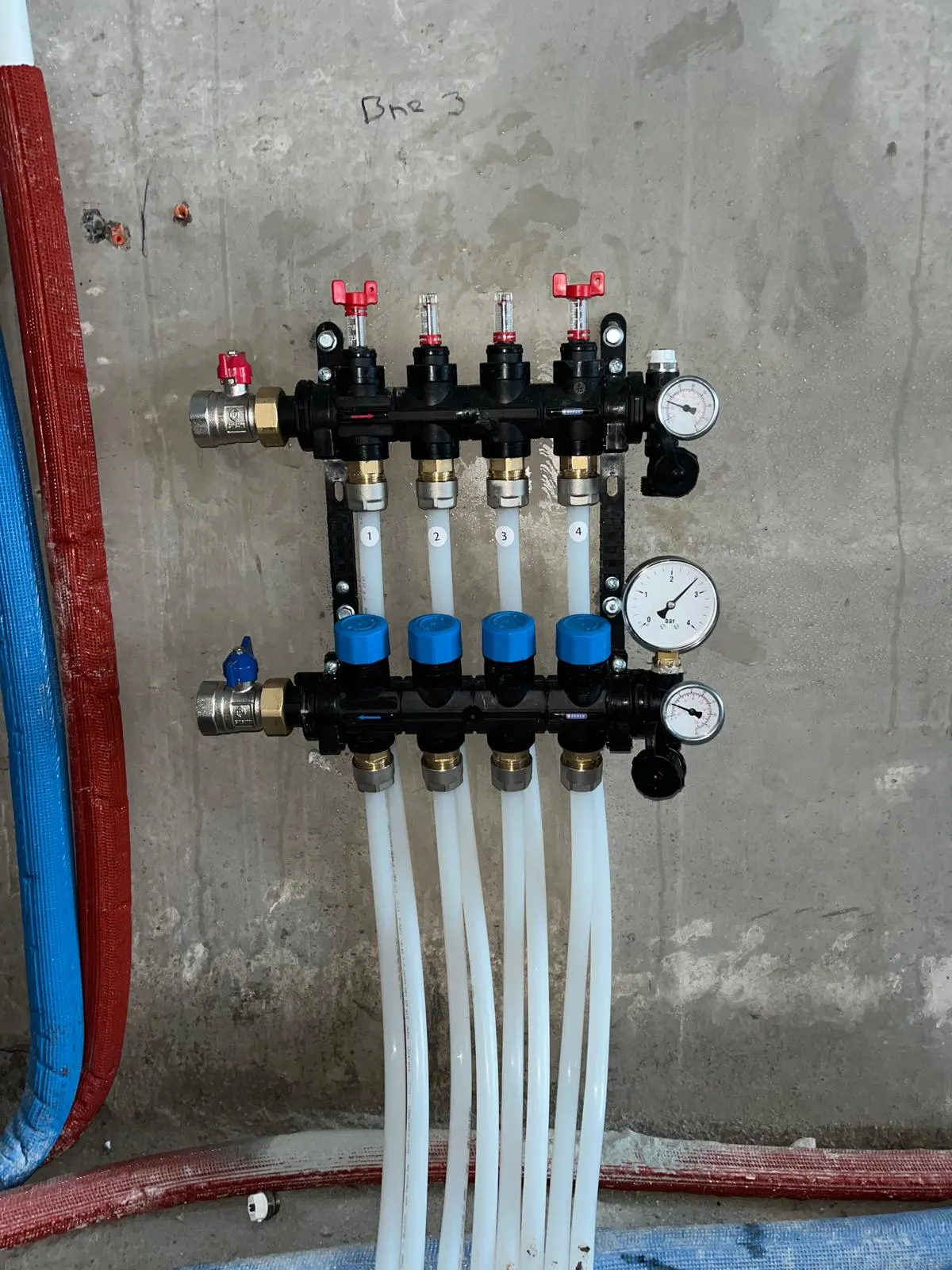 Een verdeler met vier groepen op een betonwand, met rode en blauwe afsluiters, twee manometers, genummerde doorstroommeters en witte leidingen die naar beneden de vloer in lopen