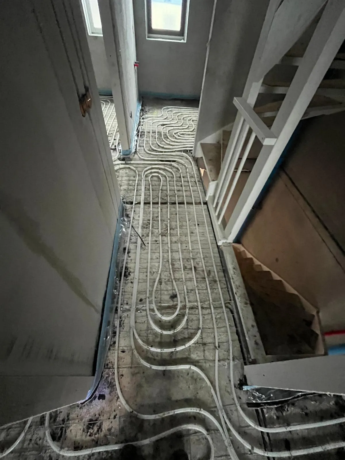 Vloerverwarmingsbuis in een lange lus op een wapeningsnet over een overloop, langs een trapgat met een wit hekwerk en houten treden, met daglicht uit een raam aan het eind