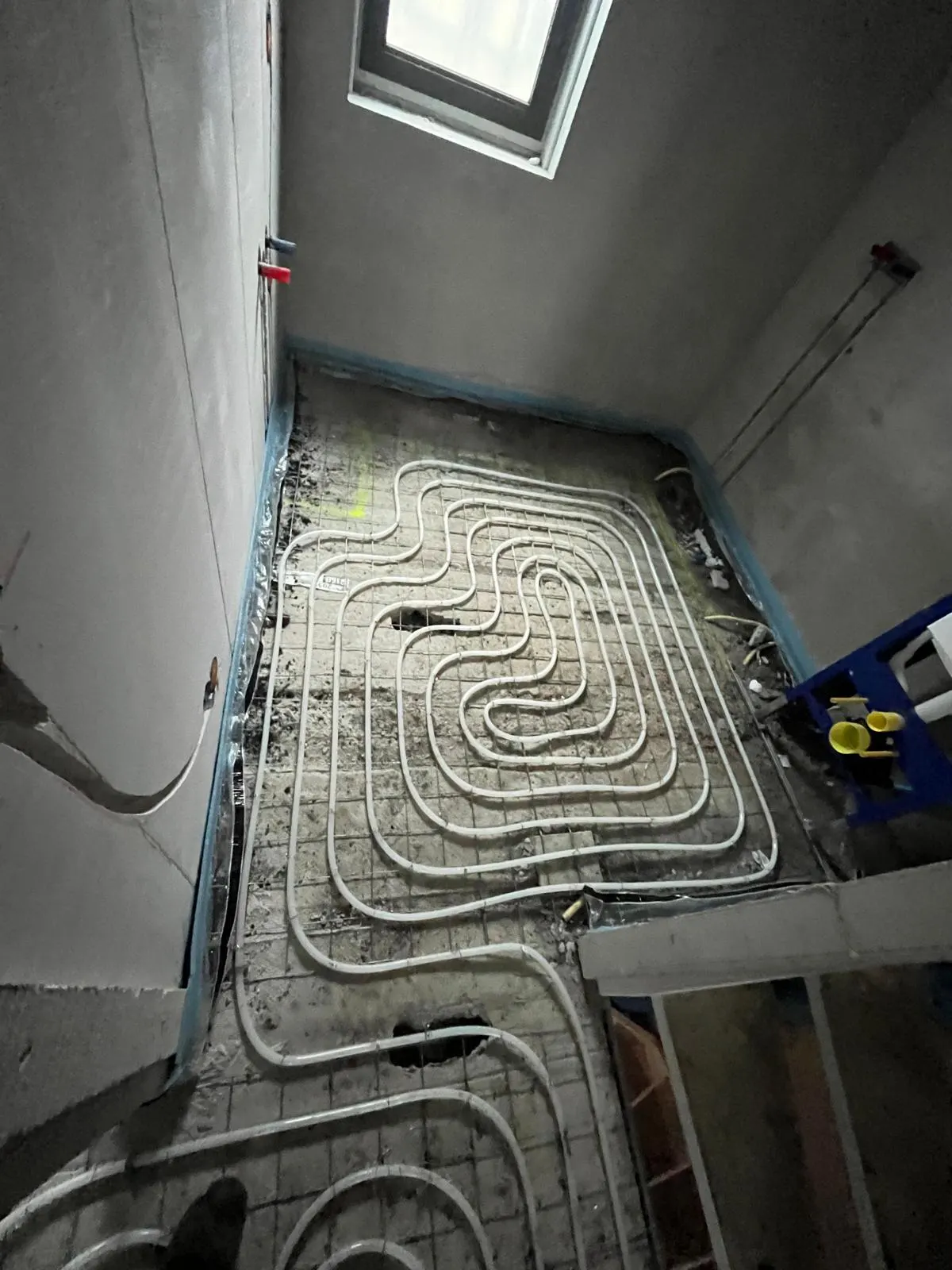 Vloerverwarmingsbuis in concentrische lussen op een wapeningsnet in een kamer met een hoog raam, met blauwe randisolatie rondom en een blauw sanitair montageframe in de hoek