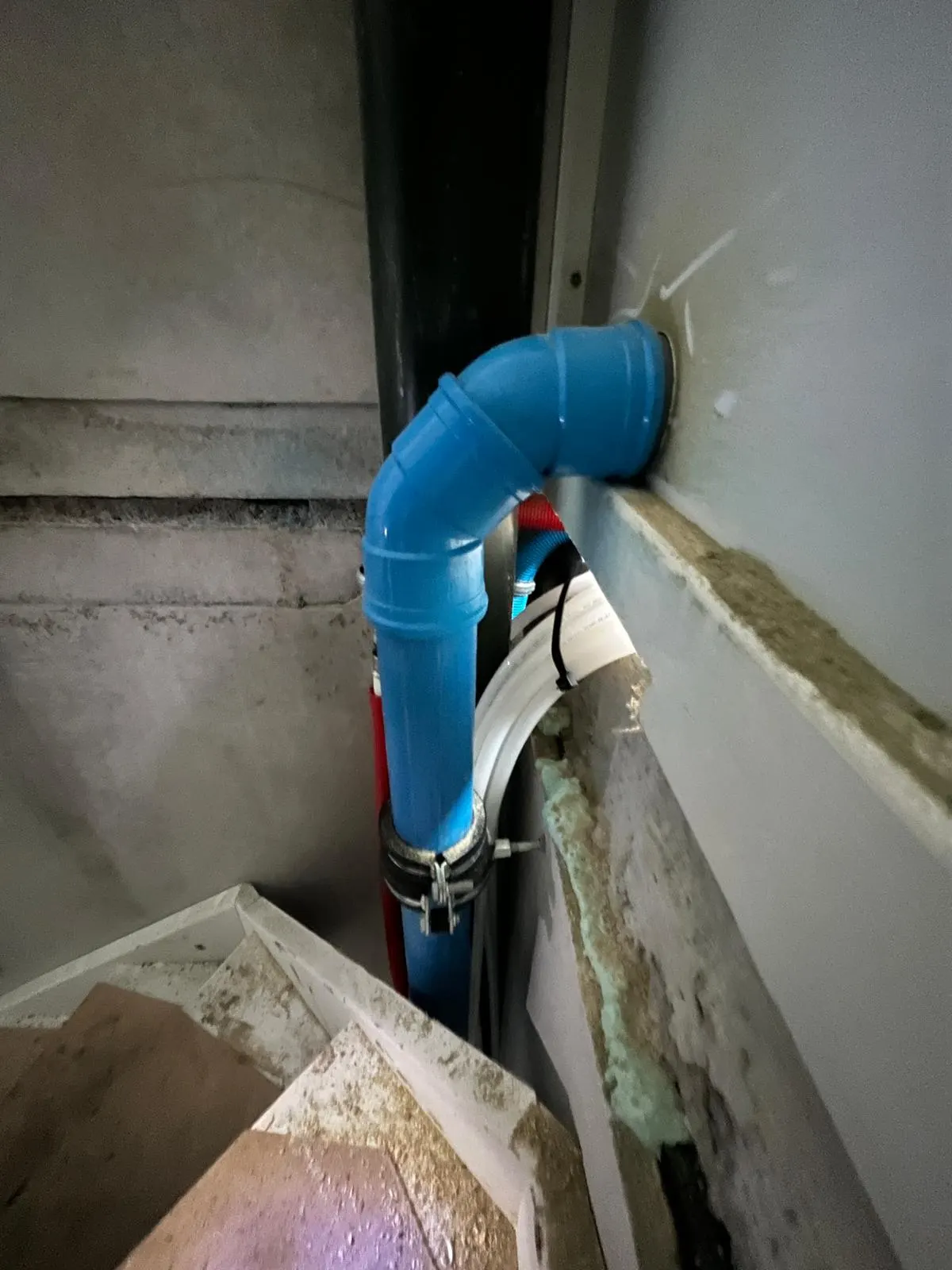 Een blauwe standleiding met een bocht die door de wand gaat, in een schacht naast een witte vloerverwarmingsleiding en een rode leiding, met de aansluiting in beeld
