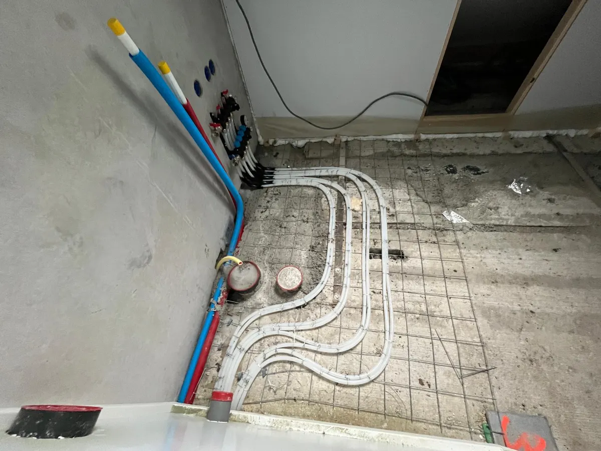 Witte vloerverwarmingsleidingen die vanaf de vloer met wapeningsnet omhoog lopen naar de aansluitpunten aan de wand, met rode en blauwe geïsoleerde leidingen langs het plafond en twee afgedopte sparingen in de vloer