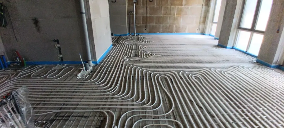 Vloerverwarmingsbuis in dichte lussen op een wapeningsnet over een hele appartementsvloer, met blauwe randisolatie langs de kalkzandsteenwanden en daglicht uit een pui op de achtergrond