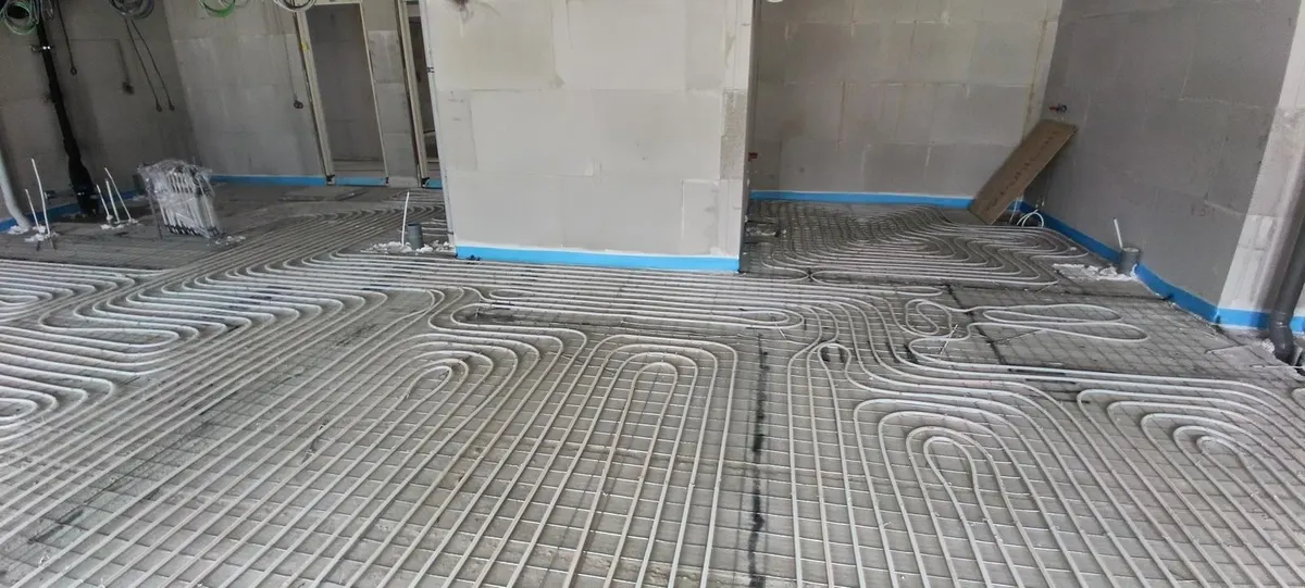 Buis in ruime lussen op een wapeningsnet in een appartement in ruwbouw, met blauwe randisolatie langs de wanden en een verdeler in beschermfolie in de hoek