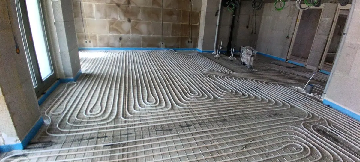 Een appartementsvloer over de volle breedte vol vloerverwarmingsbuis in dichte lussen op een wapeningsnet, met blauwe randisolatie langs de kalkzandsteenwanden en een pui met een deur links
