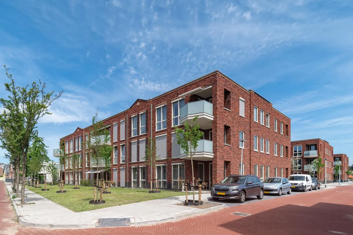 Een opgeleverd appartementengebouw van drie lagen in rode baksteen met een plat dak en een uitspringende hoek met twee balkons met matglazen borstwering, met een grasveld met jonge bomen ervoor en verderop in de straat nog twee gelijksoortige blokken