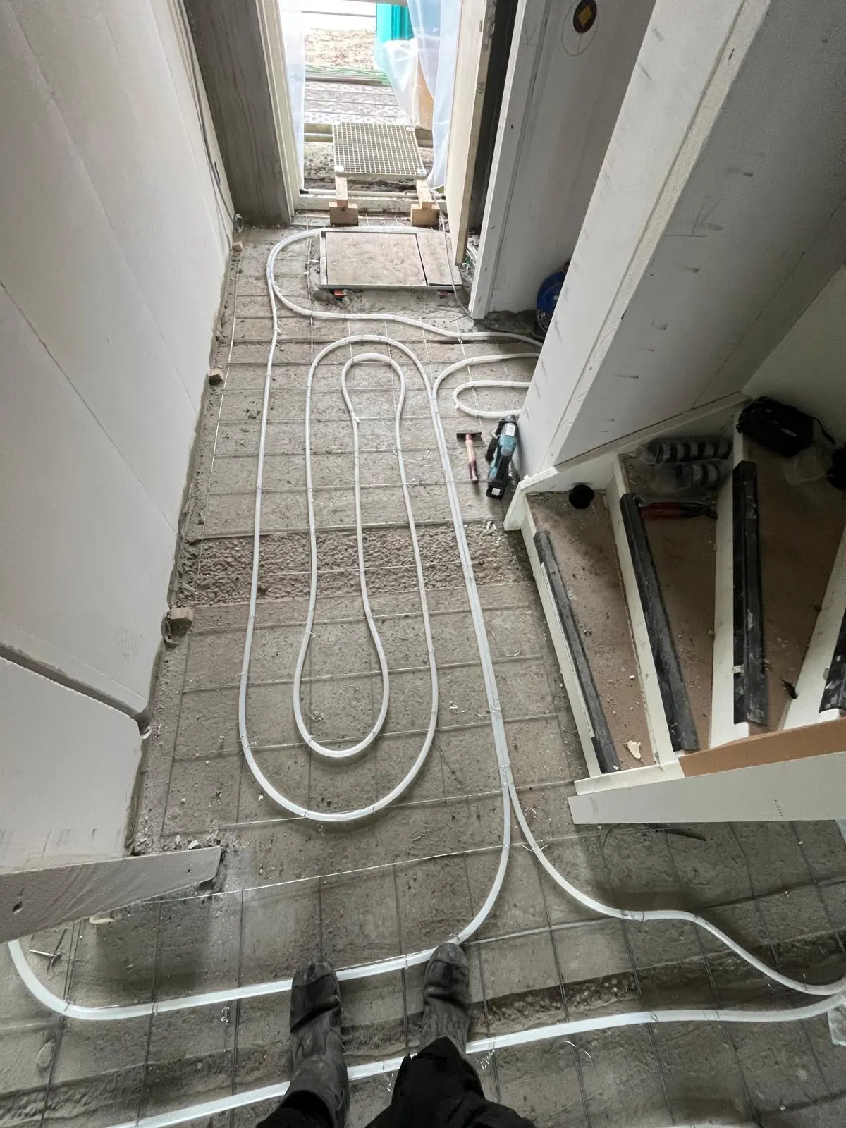 Vloerverwarmingsbuis in een lange smalle lus op een wapeningsnet in een gang, met een dichtgelegde sparing bij de voordeur, gereedschap langs de wand en de werkschoenen van de monteur onderin beeld