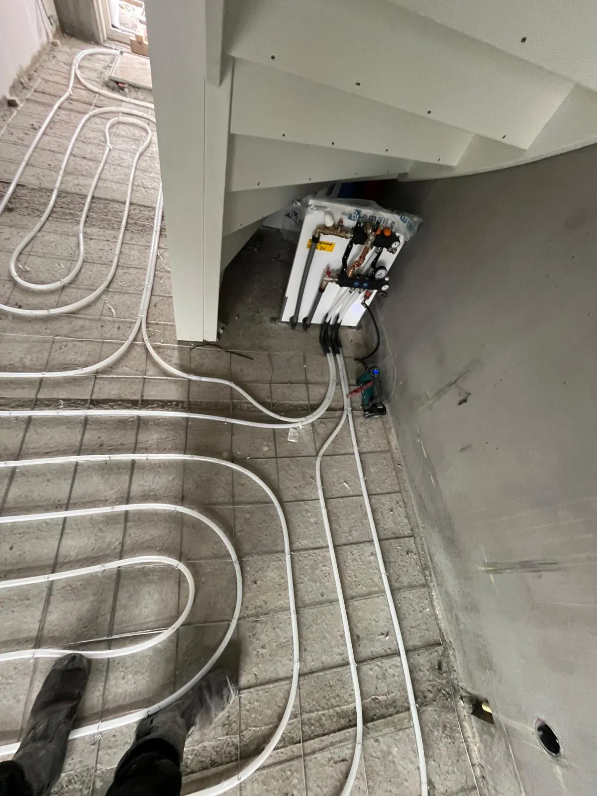 Een verdeler in beschermfolie onder een houten trap, met de aansluitleidingen die in zwarte bochtbeschermers naar de vloer lopen en vandaar over het wapeningsnet de kamer in