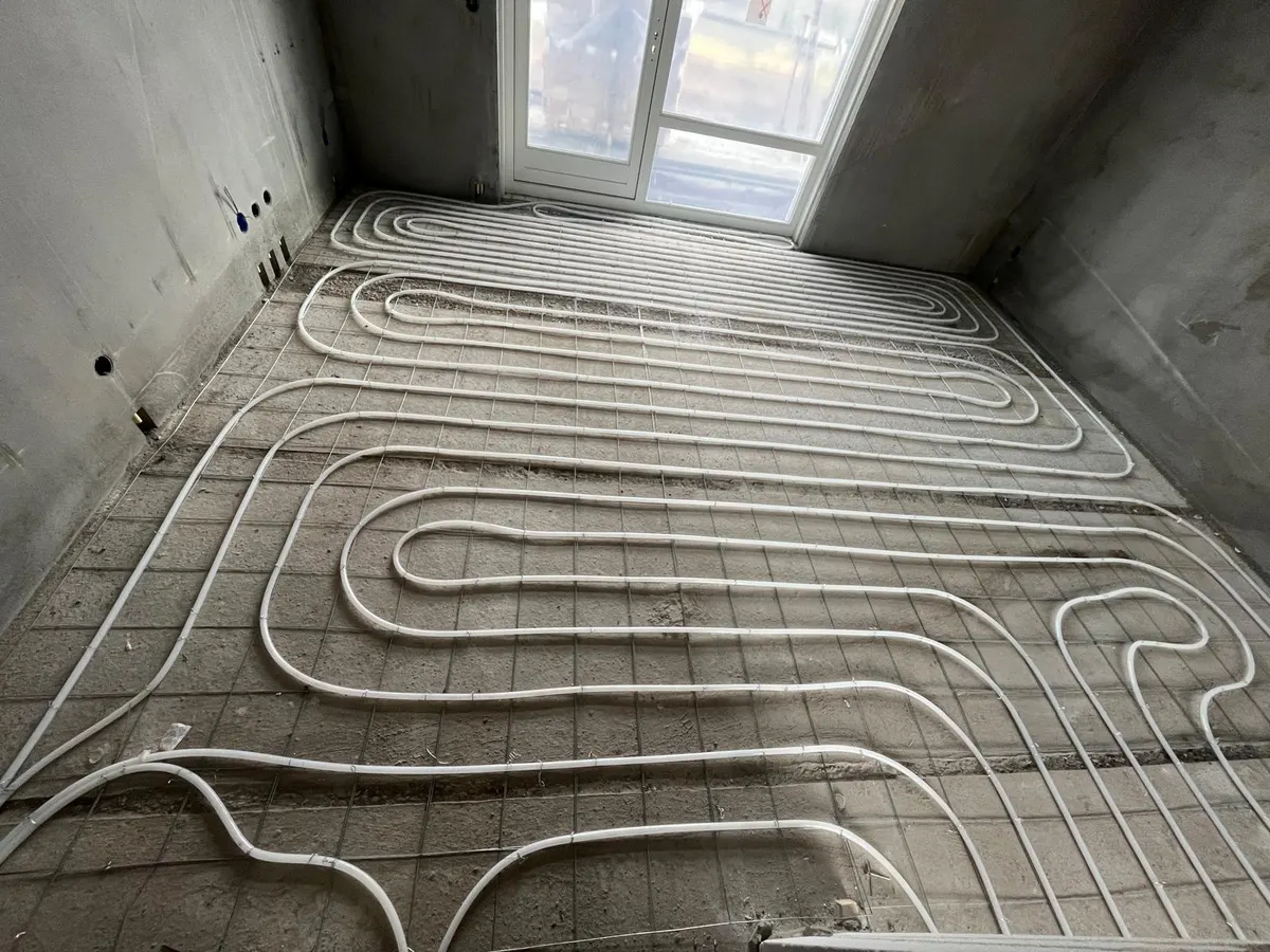 Een hele kamervloer vol vloerverwarmingsbuis in strakke lussen op een wapeningsnet, met een tuindeur met zijlicht in de achterwand en daglicht dat over de ruwe betonvloer valt
