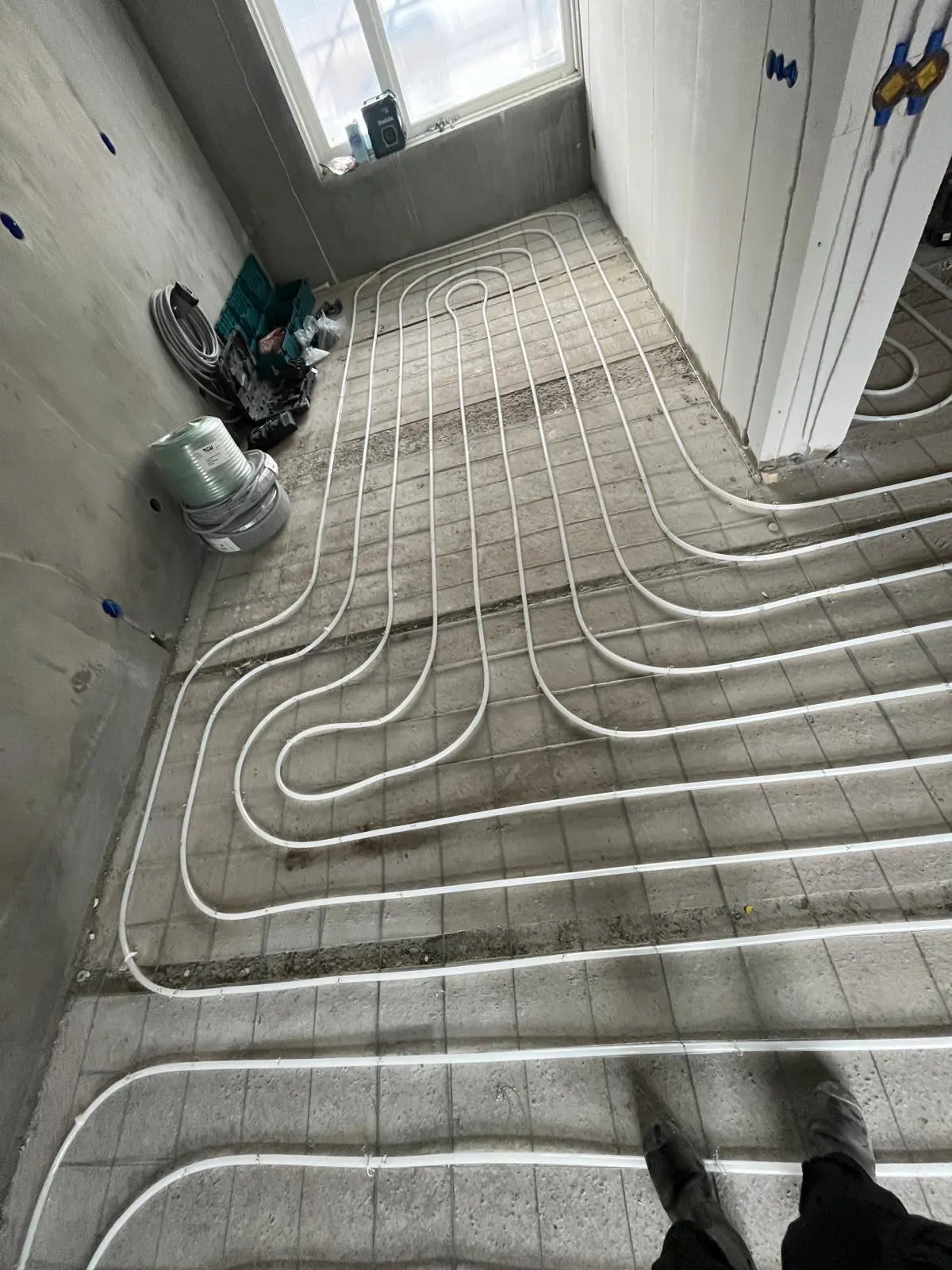 Vloerverwarmingsbuis in ineengeschoven lussen op een wapeningsnet in een kamer met een dakraam, met een rol isolatie en een gereedschapskist langs de wand