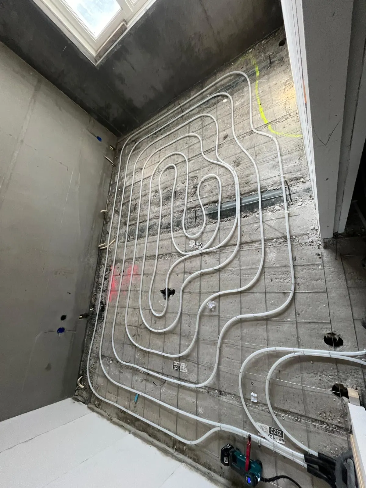 Vloerverwarmingsbuis in ronde lussen op een wapeningsnet over een betonvloer, met een dakraam boven, groepnummers op labels aan de buis en een accuschroefmachine op de vloer