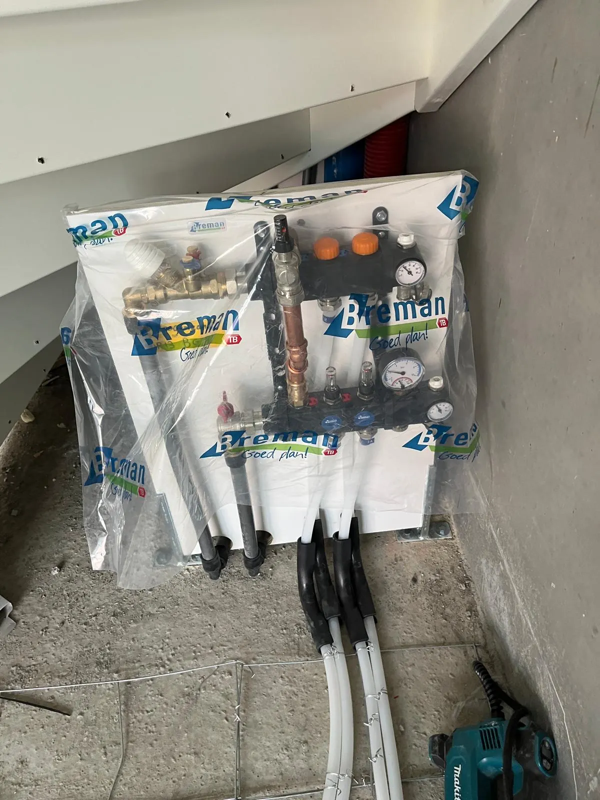 Een verdeler met twee groepen in beschermfolie op een montageplaat onder de trap, met koperen aansluitingen, twee manometers en de witte leidingen die in bochtbeschermers de vloer in gaan