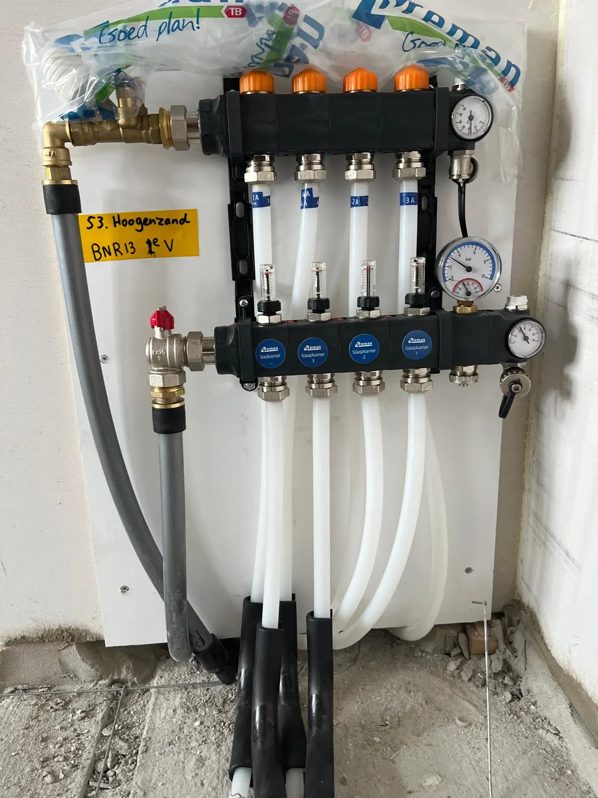 Een verdeler met vier groepen op een witte montageplaat, elke groep met een doorstroommeter en een label voor badkamer en slaapkamers, met de messing aanvoerleiding links en de witte leidingen die onderlangs in bochtbeschermers de vloer in gaan