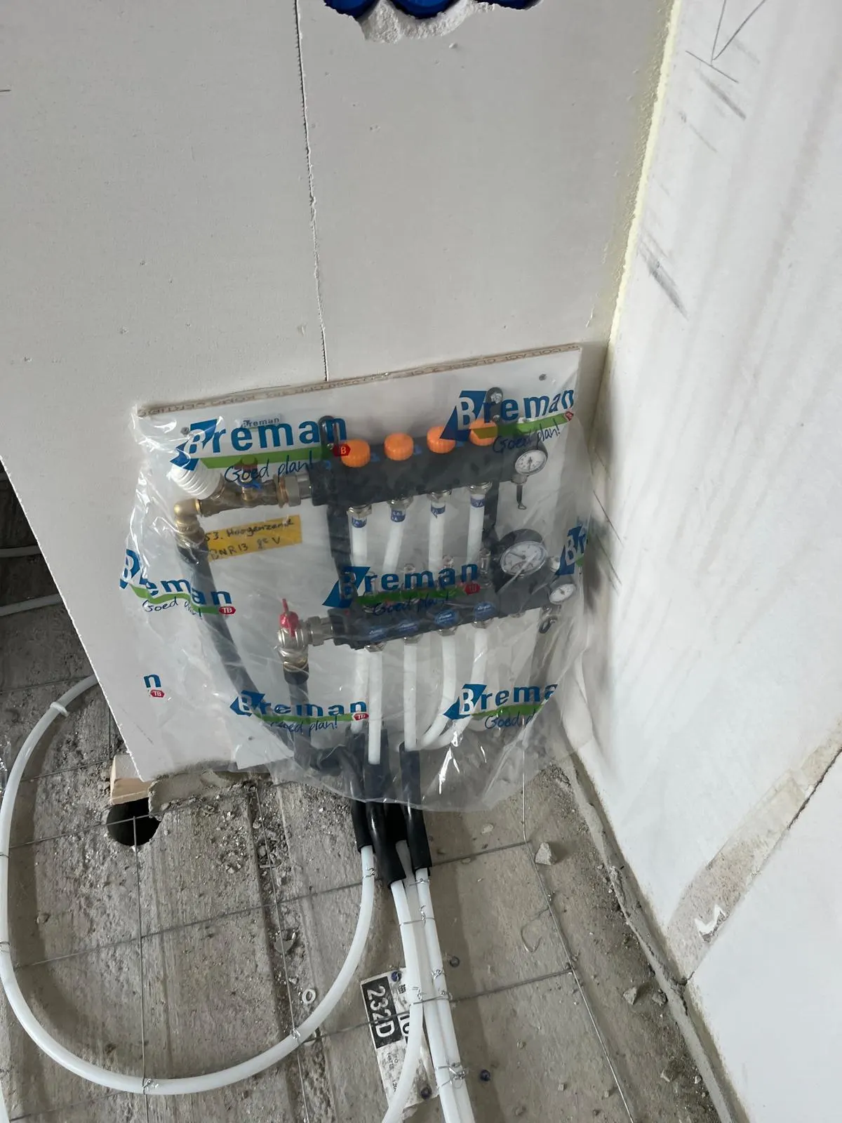 Dezelfde verdeler van bovenaf gezien, nog in beschermfolie op de plaat in de hoek van de ruimte, met de aansluitleidingen die door de bochtbeschermers over het wapeningsnet de vloer in lopen