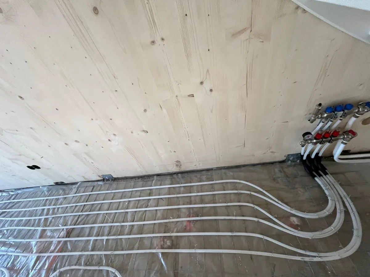 Vloerverwarmingsbuis in ruime lussen op een wapeningsnet over isolatiefolie, in een hoek van een woning met houten wanden, met de verdeler rechts aan de wand