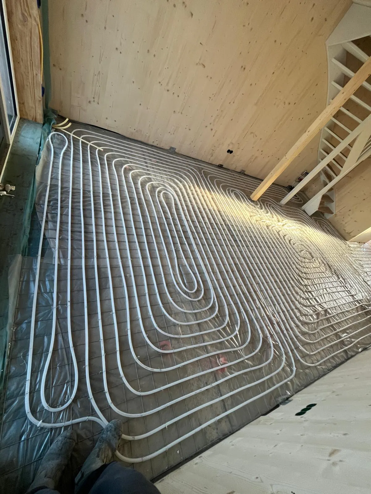 Een hele verdieping vol vloerverwarmingsbuis in lussen op een wapeningsnet, met een houten trap en houten wanden eromheen en de werkschoenen van de fotograaf onderin beeld