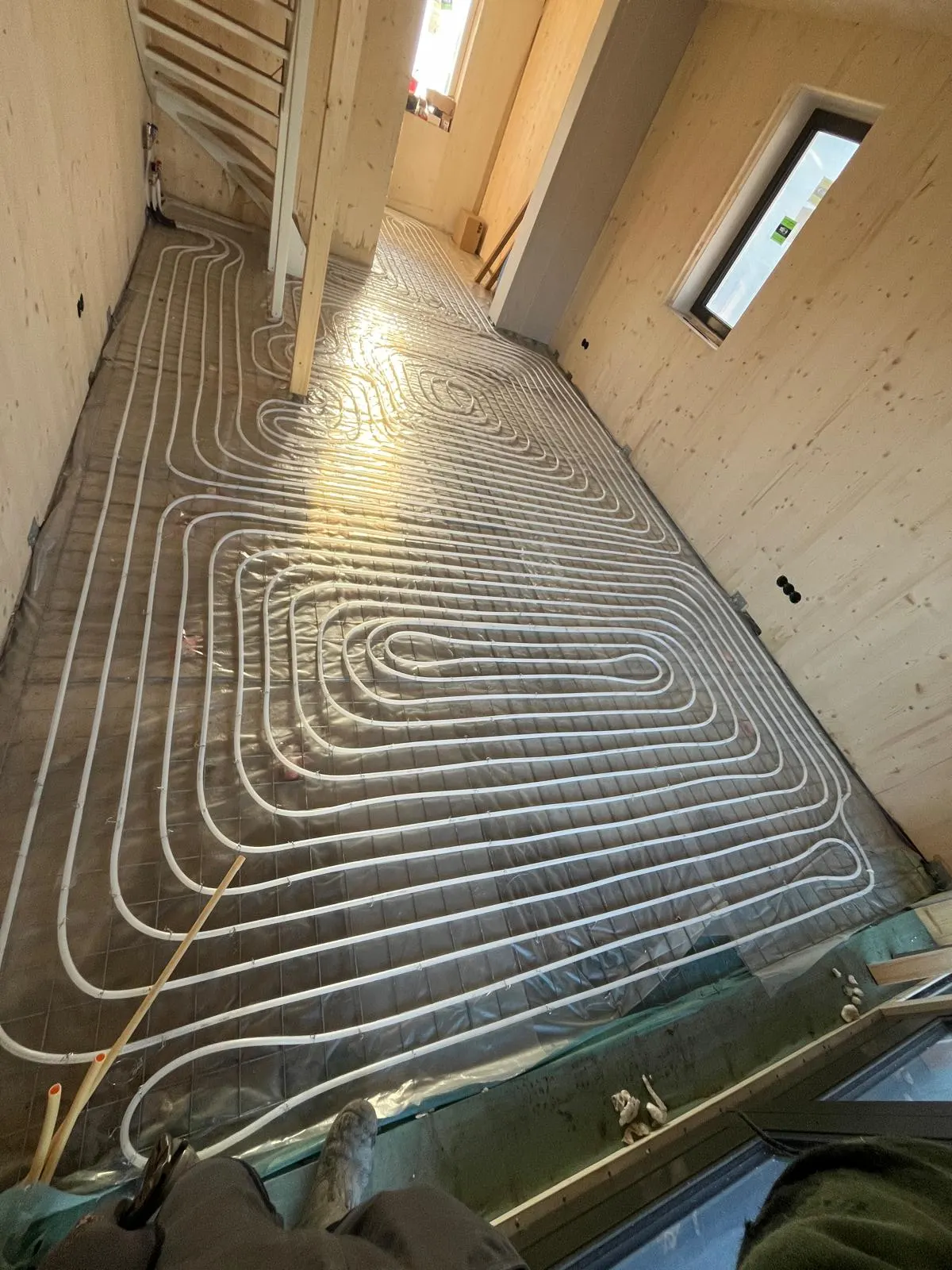 Een langgerekte verdieping vol vloerverwarmingsbuis in dichte lussen op een wapeningsnet over isolatiefolie, tussen houten wanden met een raam en een dakraam, met de werkschoenen van de fotograaf onderin beeld