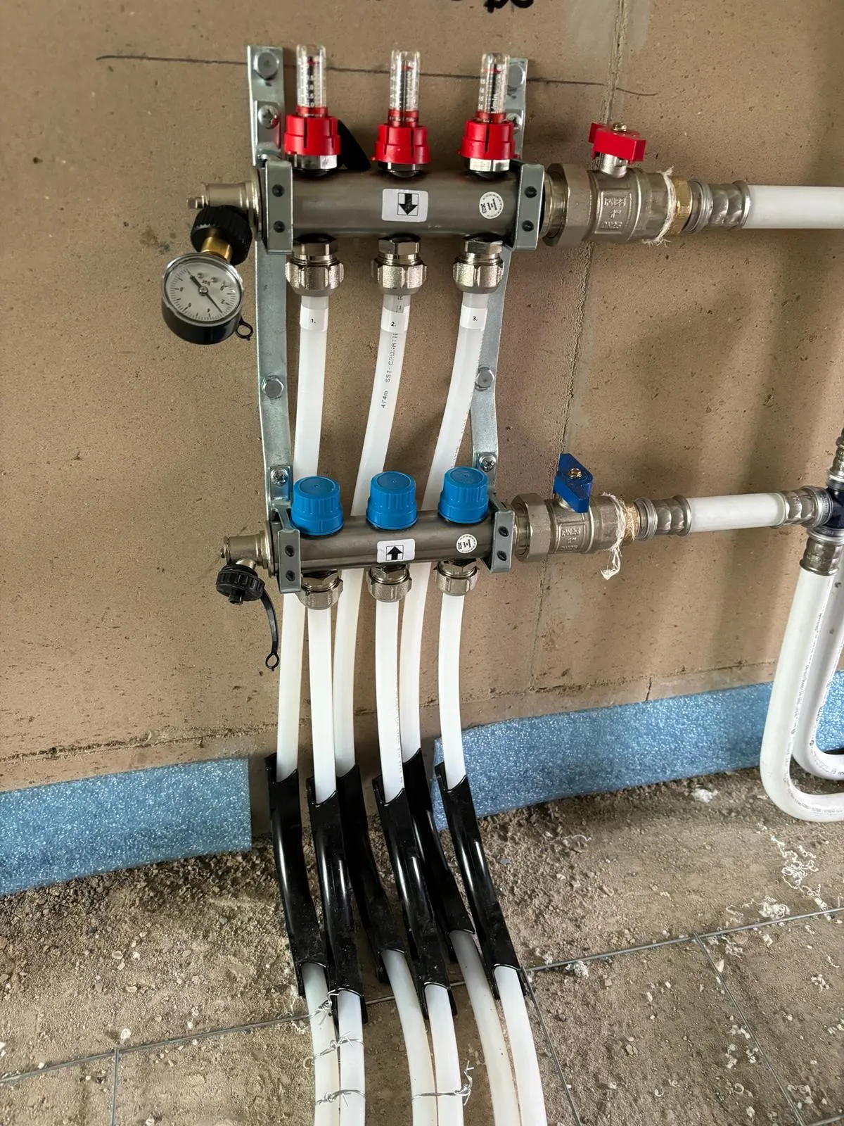 Een verdeler met drie groepen aan een wand, met de flowmeters bovenaan, rode en blauwe afsluiters en witte leidingen die in zwarte bochtbeschermers naar de vloer lopen, met blauwe randisolatie langs de wand