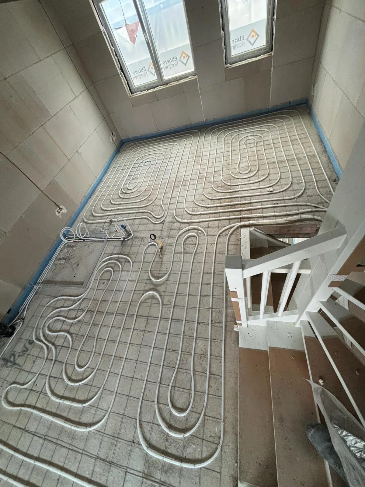 Een verdiepingsvloer vol vloerverwarmingsbuis in lussen op een wapeningsnet, met een houten trap met leuning rechts en drie ramen met beschermfolie op de achtergrond