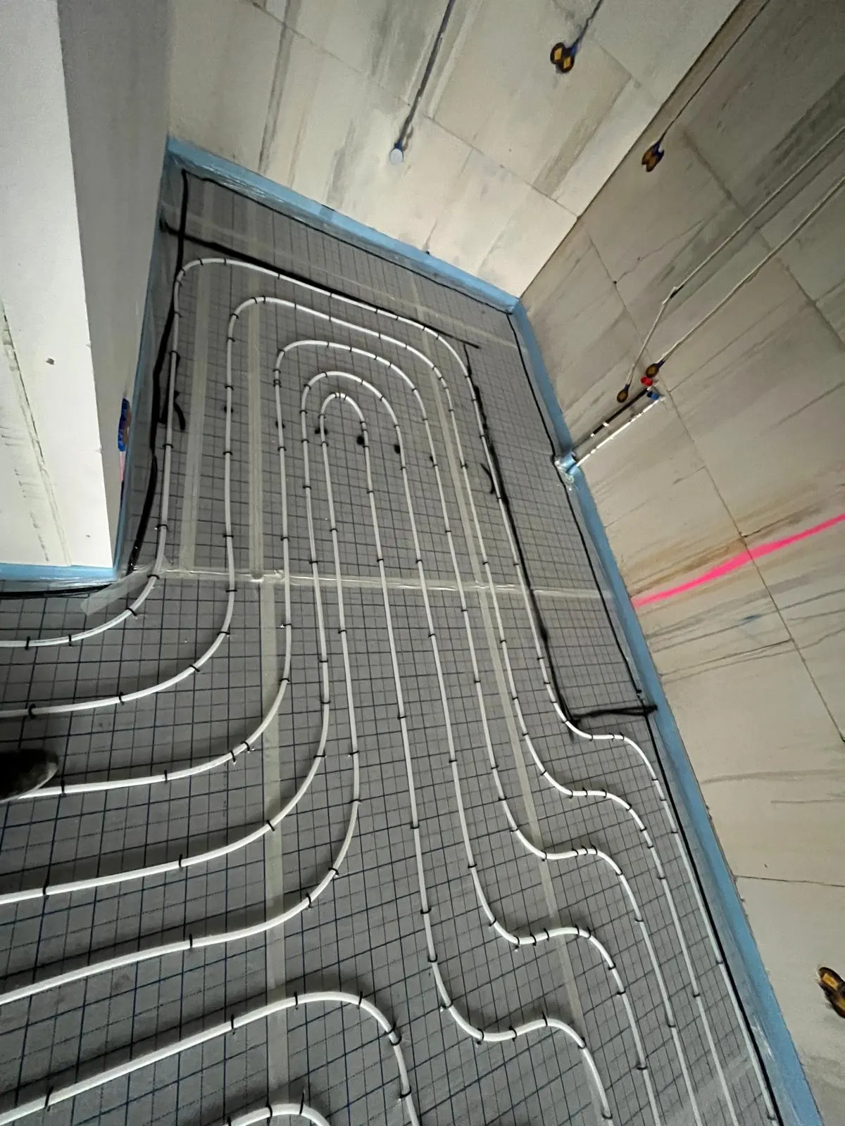Vloerverwarmingsbuis in smalle lussen op isolatieplaten in een kleine ruimte met betonnen wanden, met blauwe randisolatie rondom en een rode laserlijn op de wand ernaast