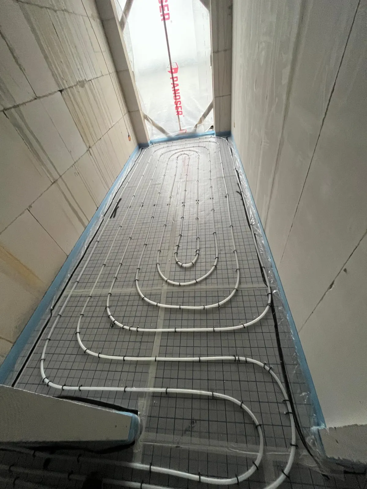 Een smalle ruimte met vloerverwarmingsbuis in een lange lus op isolatieplaten tussen twee betonwanden, met daglicht uit de opening aan het eind