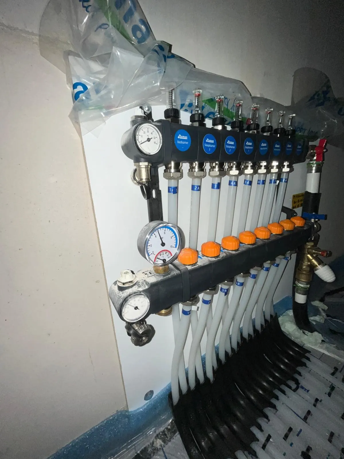 De verdeler van de woning aan de wand, met tien groepen, doorstroommeters en handwielen, en de aansluitleidingen die er in een waaier naartoe lopen