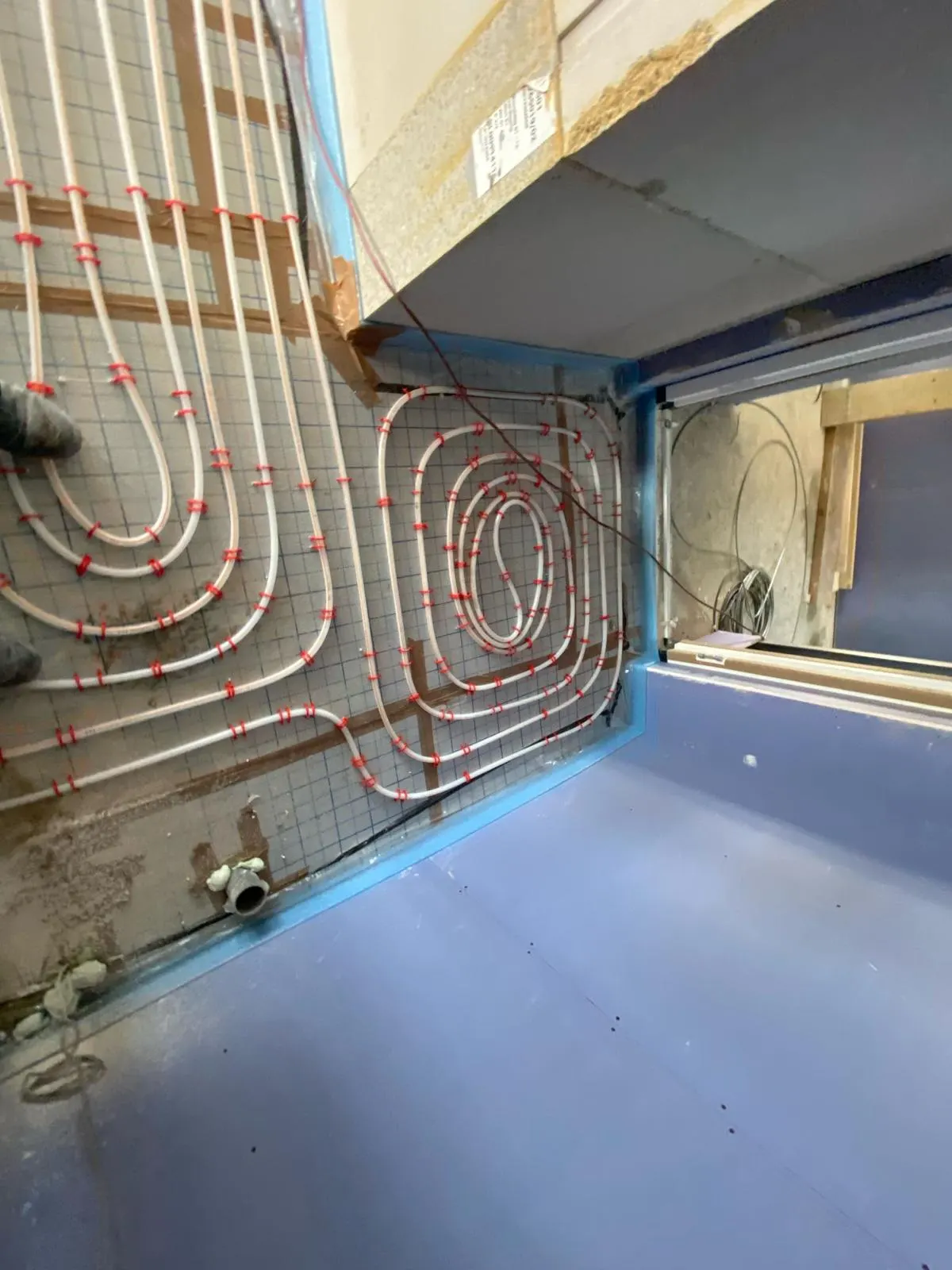Vloerverwarmingsbuis in een slakkenhuis op een wapeningsnet in een kleine ruimte, met een deuropening naar een gang met een afgewerkte vloer