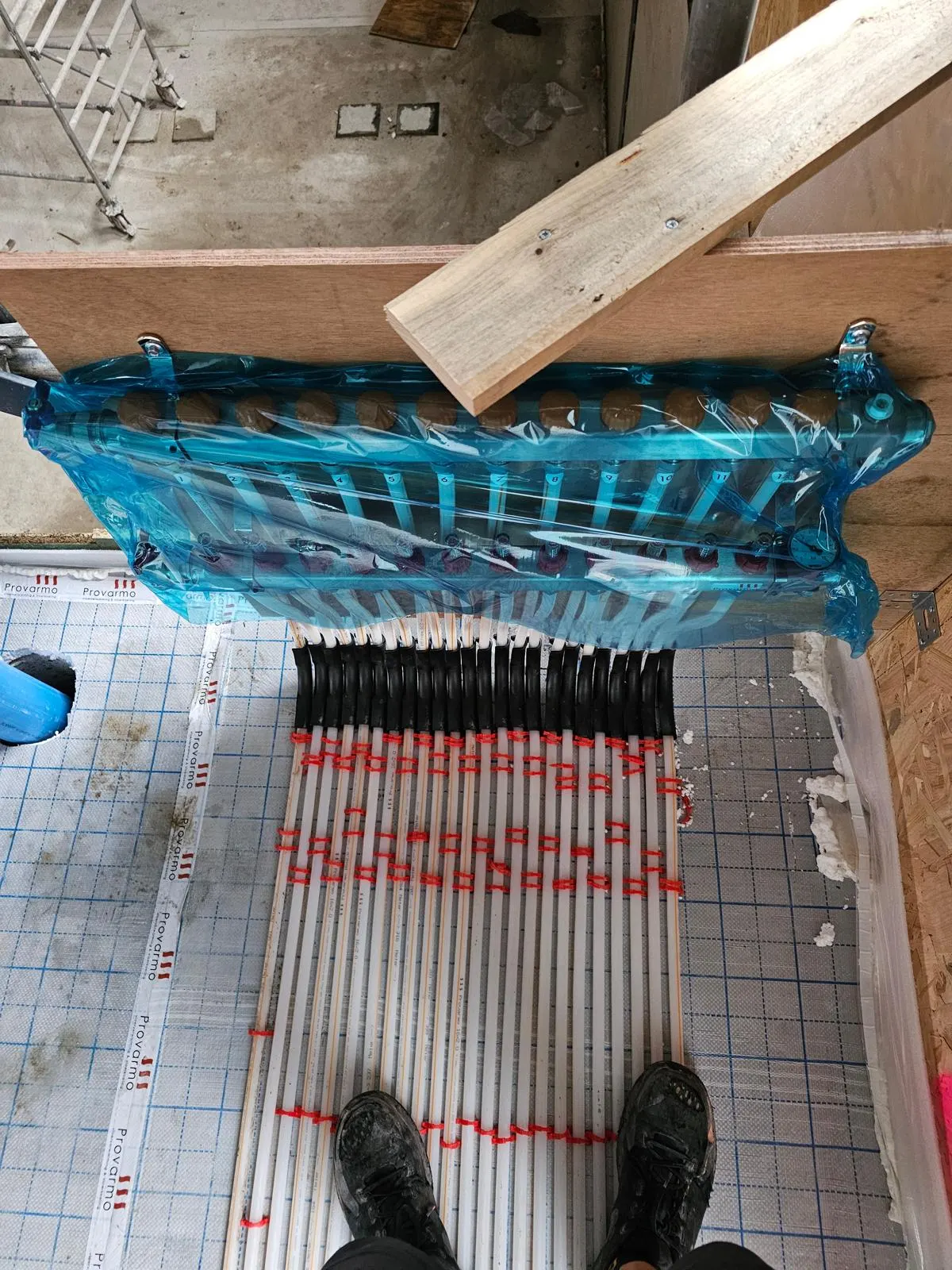 Een verdeler in blauwe beschermfolie boven een bundel vloerverwarmingsbuizen die met rode clips op tackerfolie naar hem toe lopen, met een houten bouwtrap ernaast en de werkschoenen van de fotograaf onderin beeld