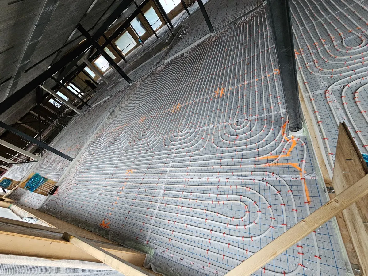 Vloerverwarmingsbuis op tackerfolie tot tegen een houten stijlwand in aanbouw, met steigerwerk in de ruimte erachter