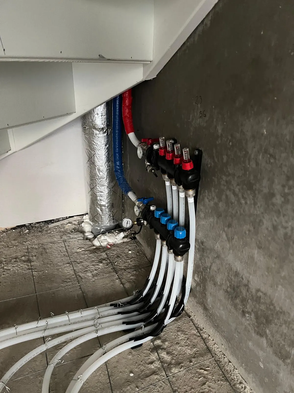 Een verdeler met vijf groepen laag tegen een betonwand in een hoek, met rode en blauwe afsluiters, een manometer en witte vloerverwarmingsleidingen die in een ruime bocht over het wapeningsnet wegdraaien