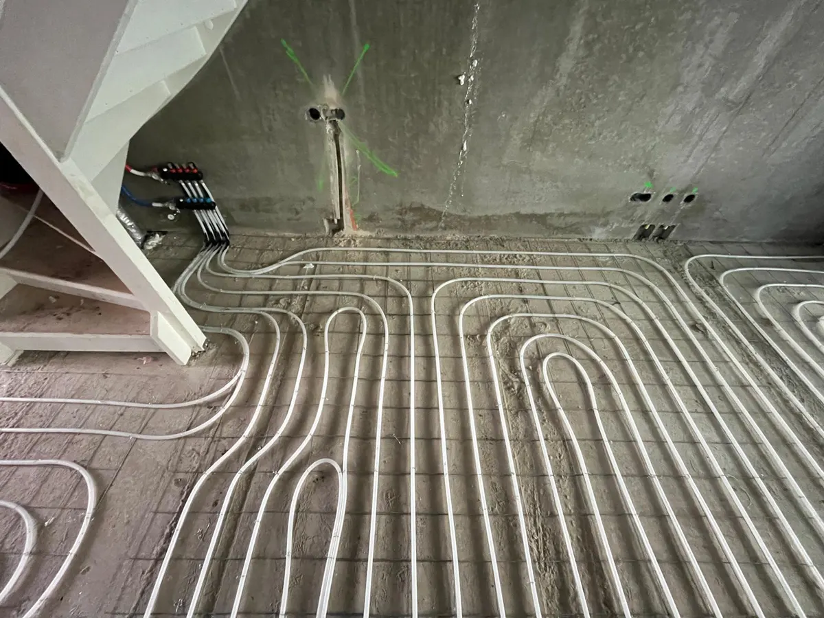 Vloerverwarmingsbuis in strakke lussen op een wapeningsnet over een kamervloer, met een verdeler tegen de wand onder een trap en groene markeringen rond een leidingdoorvoer