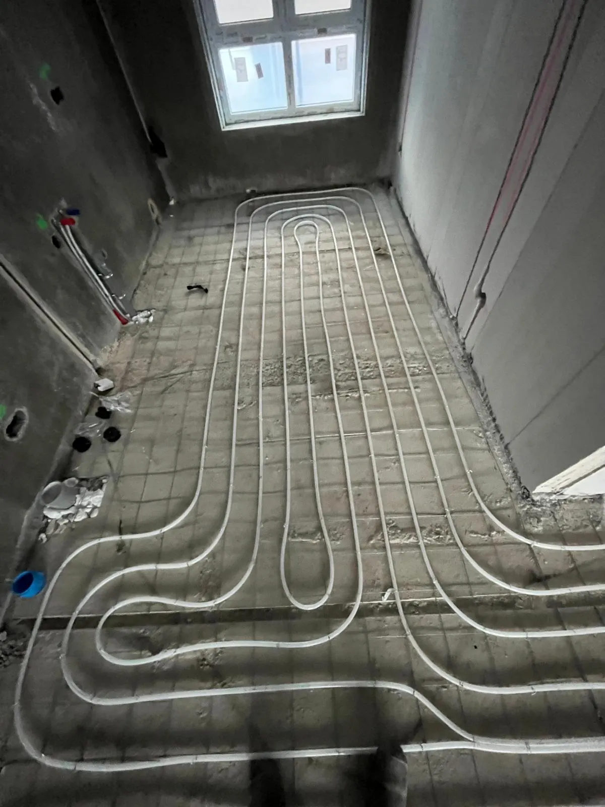 Vloerverwarmingsbuis in een enkele lus op een wapeningsnet in een smalle kamer met een hoog raam, met de aansluitpunten voor water en elektra tegen de linkerwand