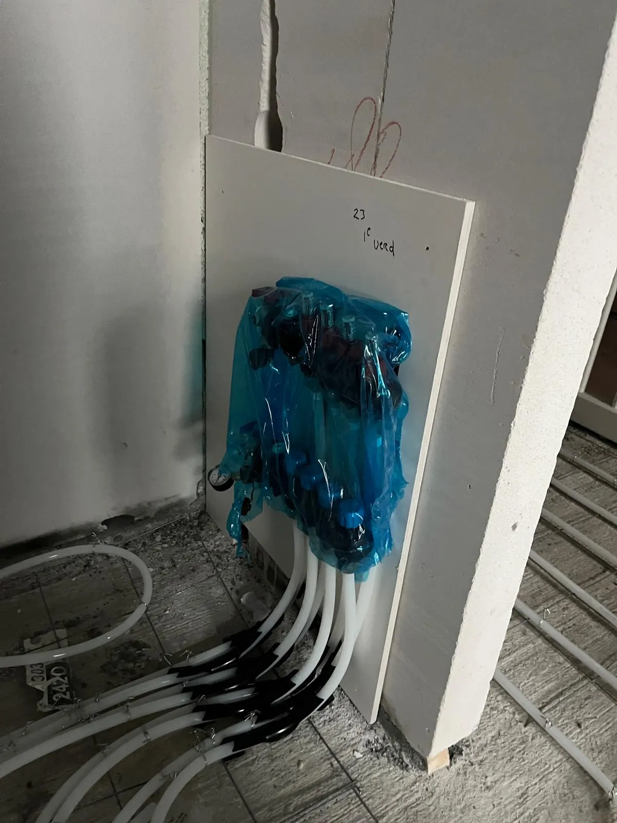 Een verdeler in blauwe beschermfolie op een montageplaat tegen een gipsplaatwand, met witte leidingen die er onderuit komen en over de vloer weglopen