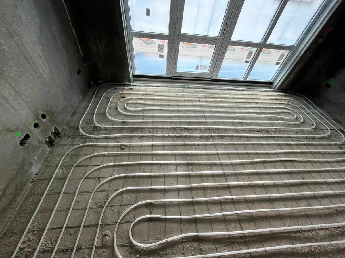 Vloerverwarmingsbuis in ineengeschoven lussen op een wapeningsnet over een kamervloer, met een brede raampui met daglicht aan de kopse kant en groene markeringen op de betonwand