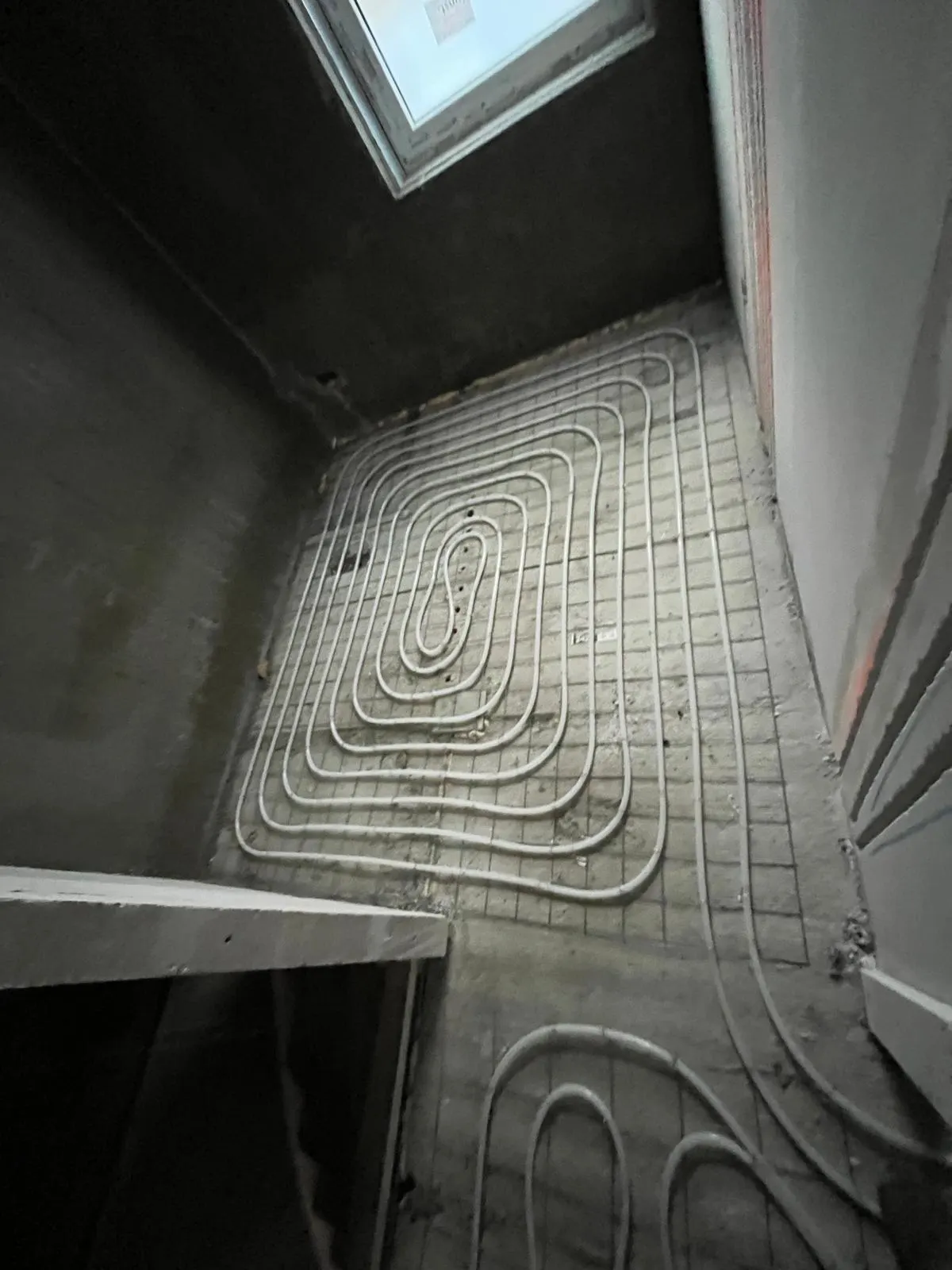 Een slakkenhuis van vloerverwarmingsbuis op een wapeningsnet in een kamer met een schuin raam, met de aanvoer die vanaf de doorgang de ruimte in loopt