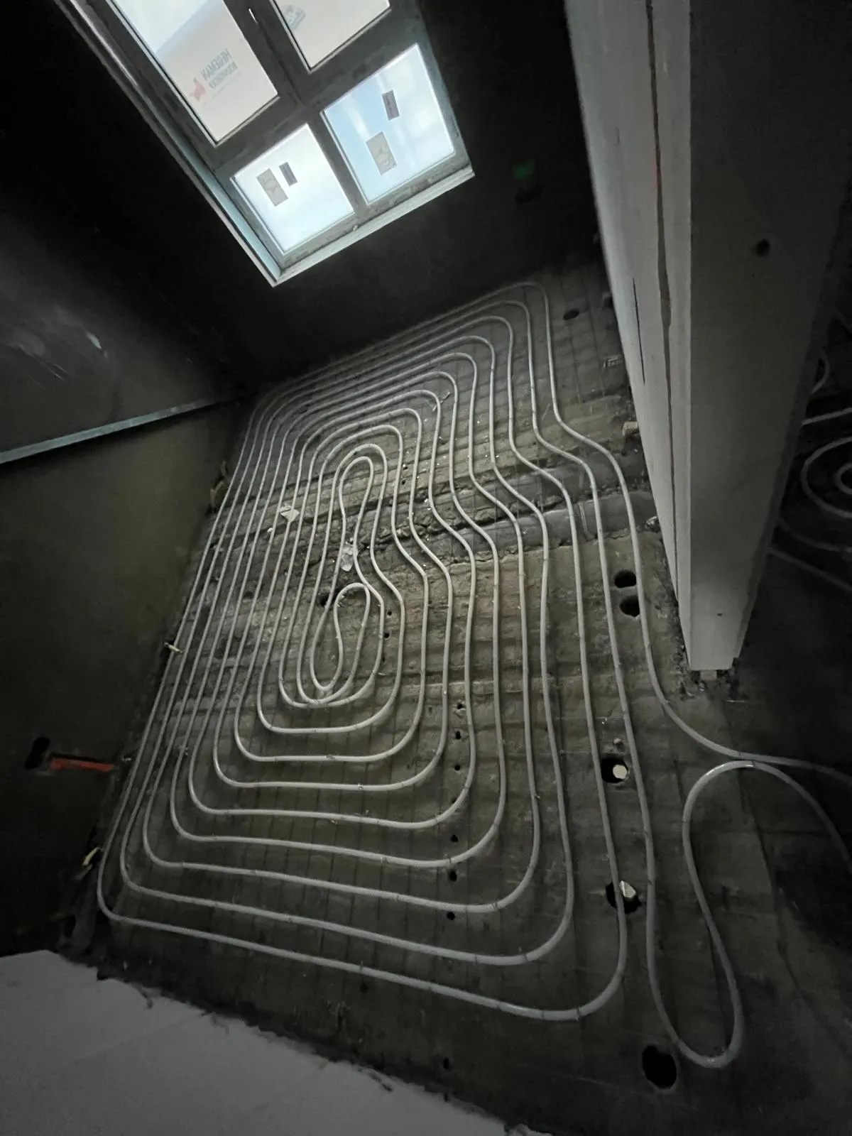 Vloerverwarmingsbuis in een groot slakkenhuis op een wapeningsnet over een kamervloer, met een raam met daglicht boven en een gipsplaatwand met sparingen aan de zijkant