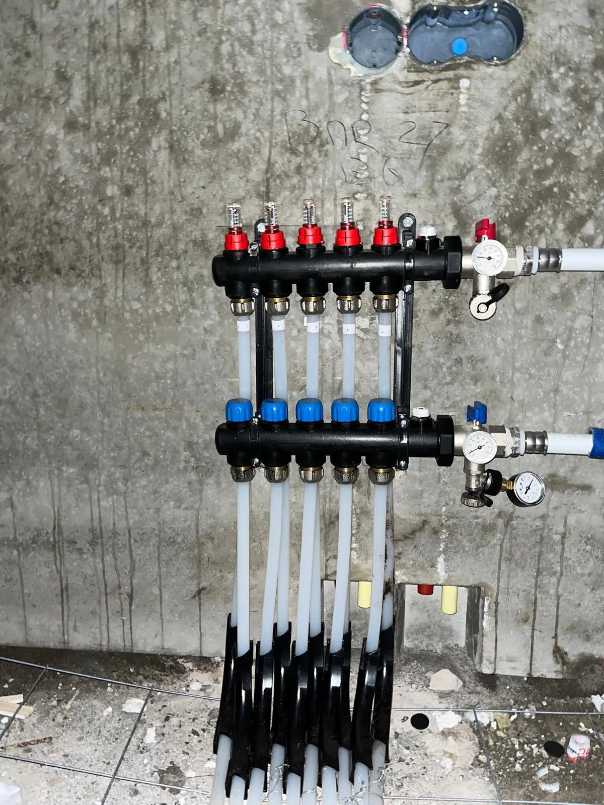 Een verdeler met vijf groepen tegen een betonwand, met genummerde doorstroommeters, rode en blauwe afsluiters, twee manometers en witte leidingen die naar de vloer lopen