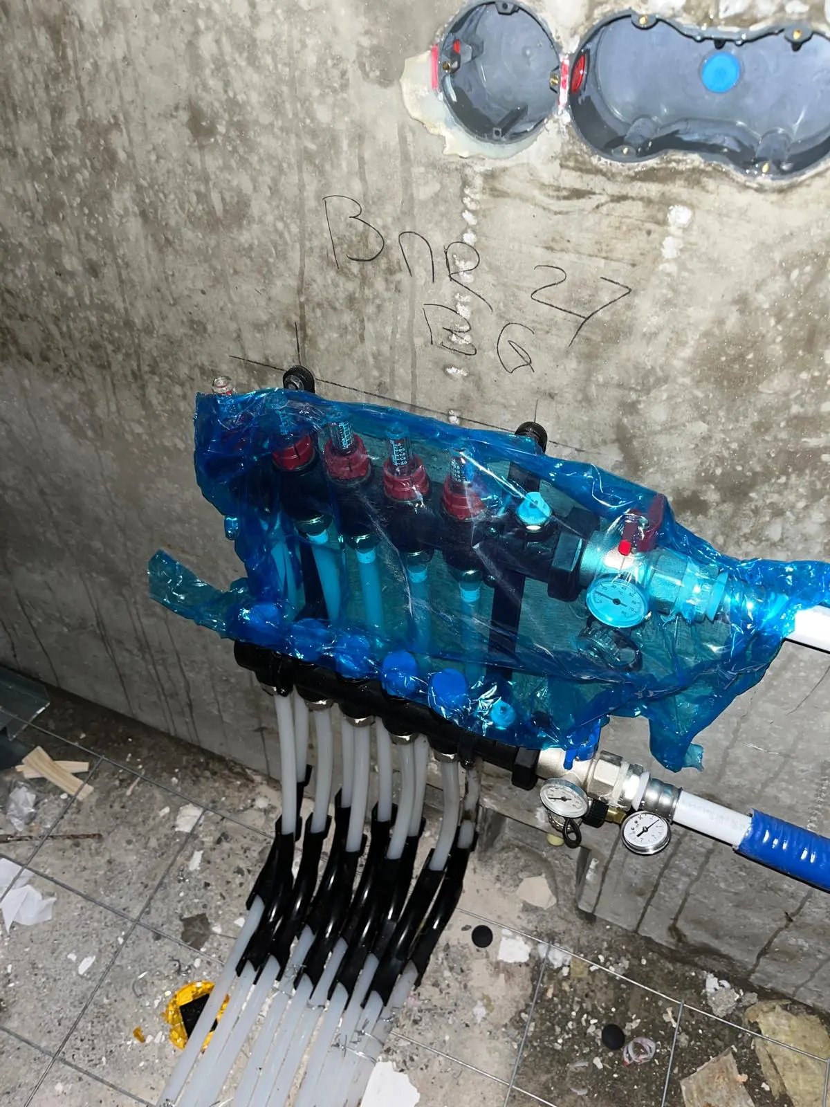 Een verdeler in blauwe beschermfolie tegen een betonwand met een krijtaanduiding erboven, met de doorstroommeters en de manometers door de folie heen zichtbaar