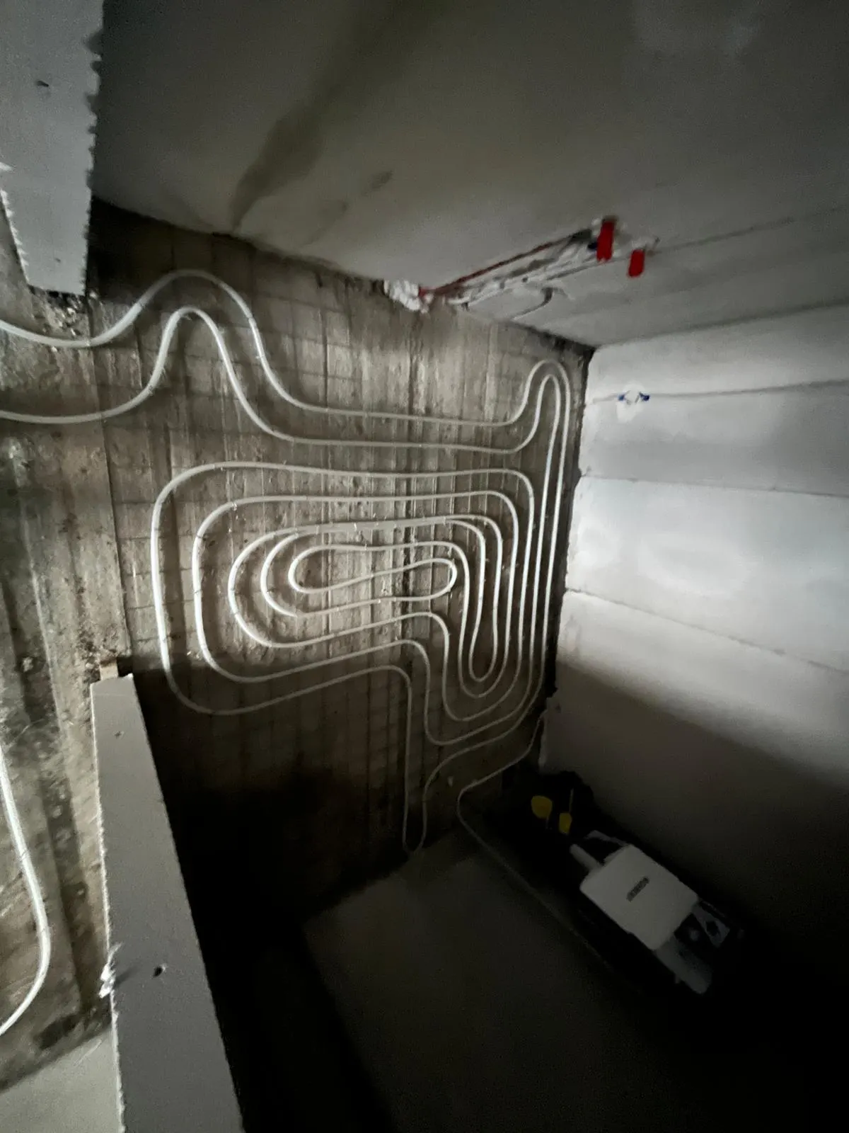 Vloerverwarmingsbuis in ineengeschoven lussen op een wapeningsnet tegen een ruwe betonwand, met een geïsoleerde leiding langs het plafond en een installatiekast in de hoek